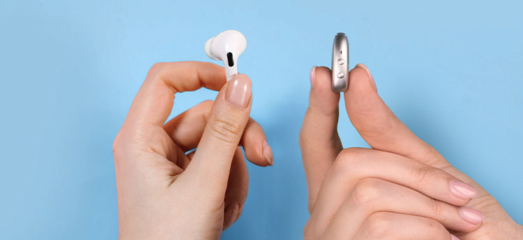 Zwei Hände halten ein Hörgerät und einen Apple AirPod 2 vor einem blauen Hintergrund.